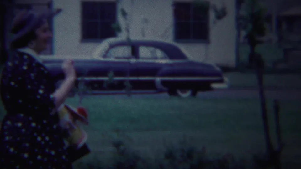 A woman in 1950s dress and hat walks through a residential outdoor scene in South Bend, with a dark sedan and low building behind her.