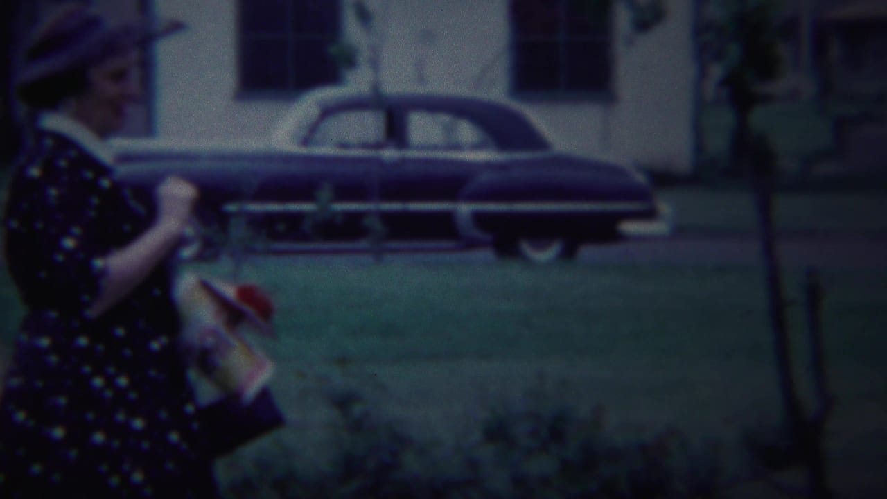 In South Bend, Indiana, a woman in 1950s dress and hat walks along a residential sidewalk with parked sedans in the background.