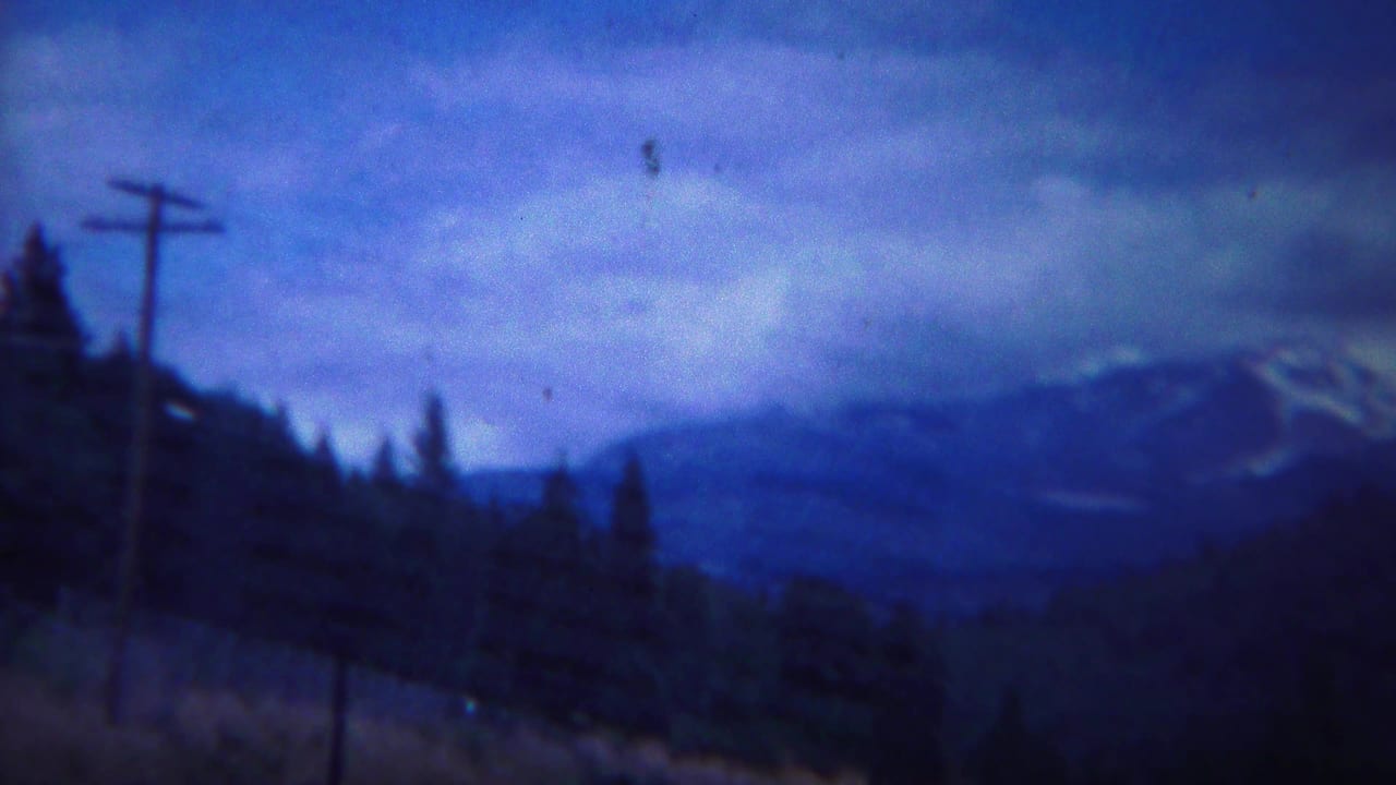 1967 8mm Yellowstone-area mountain view with a utility pole in the foreground, dark forest, cloudy blue sky, and distant snow-capped ridges.