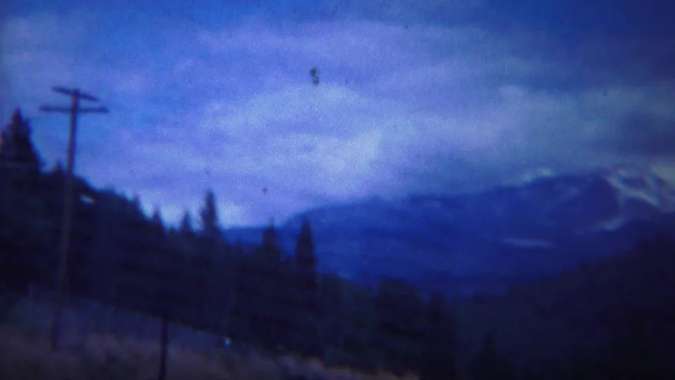 1967 8mm Yellowstone-area mountain view with a utility pole in the foreground, dark forest, cloudy blue sky, and distant snow-capped ridges.