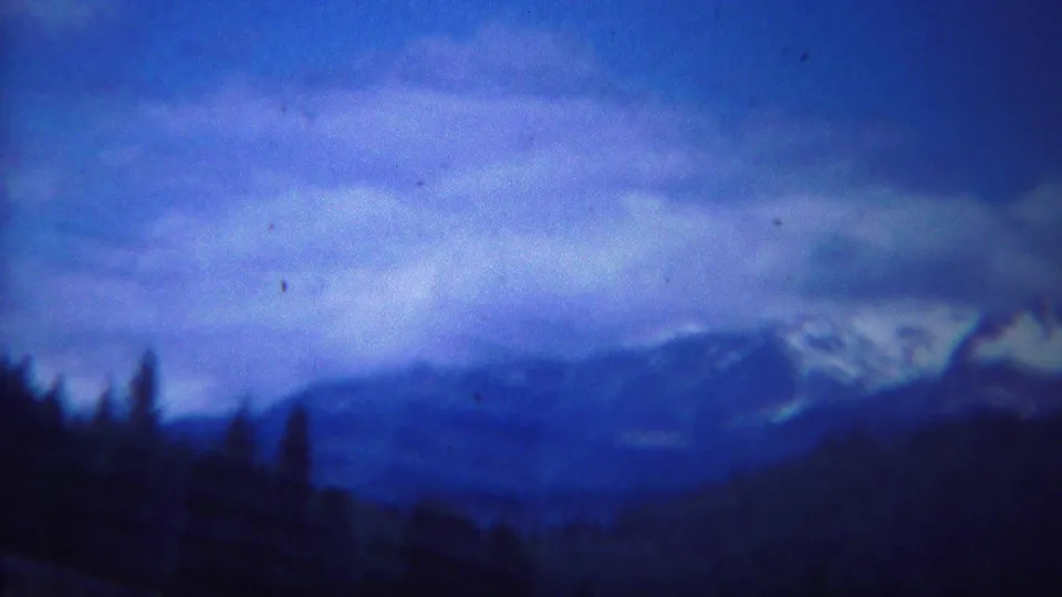 Blue 8mm frame of Yellowstone-area mountains and forested slopes under a broad cloud layer, with soft focus and film grain.