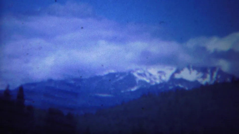 1967 8mm Yellowstone-area view of a snow-streaked ridge, dark forest foreground, and thick blue cloud cover.