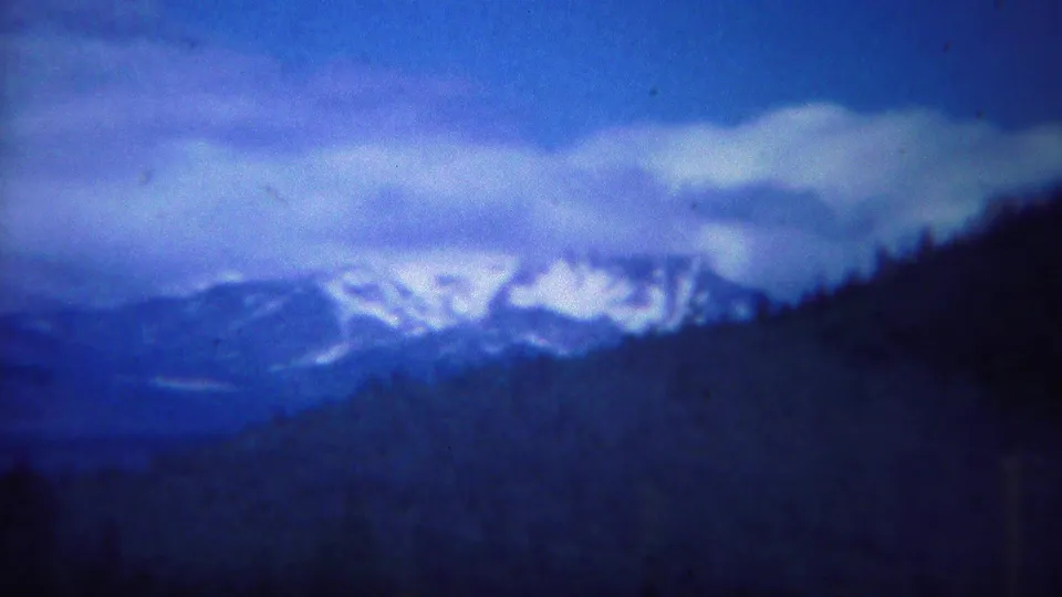Yellowstone-area 8mm landscape frame showing distant snow-capped mountains, dark forested slopes, and a heavy blue cloud bank.