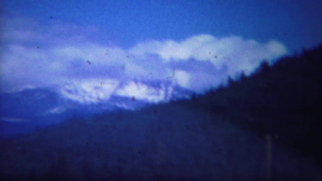 1967 Yellowstone home-movie frame showing snowy mountain ridges beyond forested hills beneath a deep blue, clouded sky.