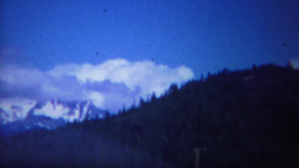 1967 Yellowstone frame with a dark tree-covered ridge crossing the foreground and snow-bright mountains partly hidden by clouds.