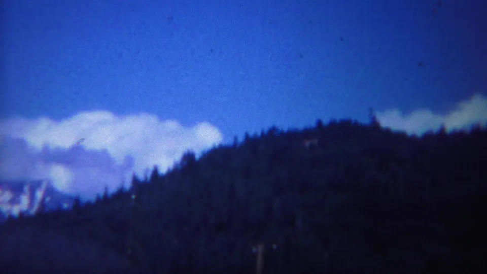 1967 Yellowstone home-movie view of clouds and a partially visible snowy mountain beside a dark wooded hillside.
