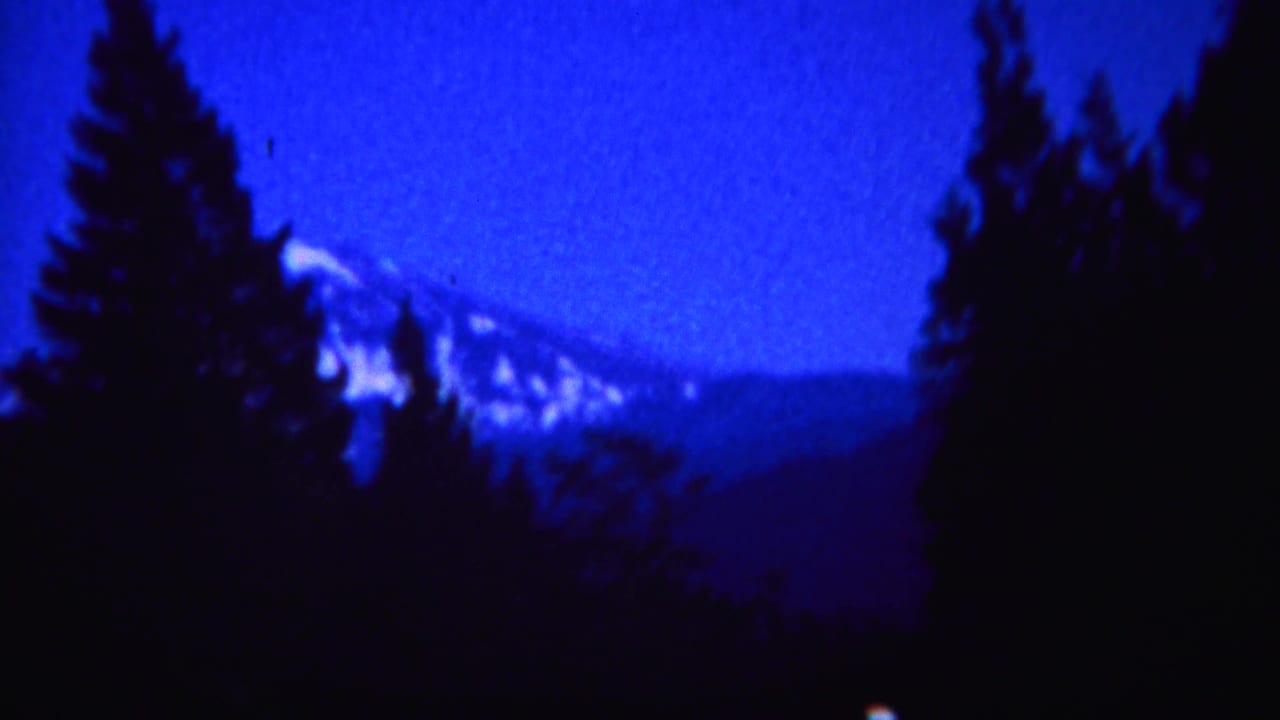 1967 Yellowstone home-movie frame with pine silhouettes and a distant snow-capped mountain in blue low light.