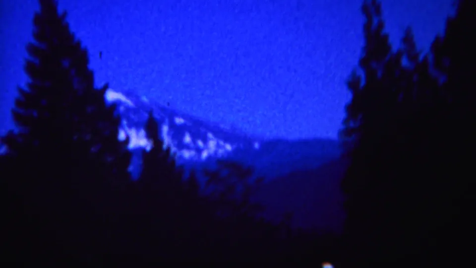 1967 Yellowstone home-movie frame with pine silhouettes and a distant snow-capped mountain in blue low light.