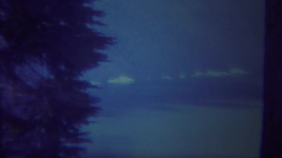 1967 Yellowstone-area 8mm frame with a dark evergreen edge, blue twilight sky, faint clouds, and a calm distant horizon.