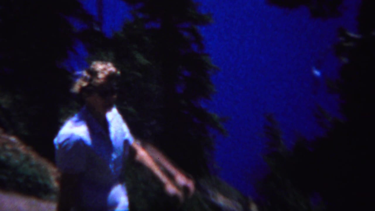 1967 Yellowstone home-movie frame showing a person in a light shirt moving past dark evergreens near a blue lakeside slope.