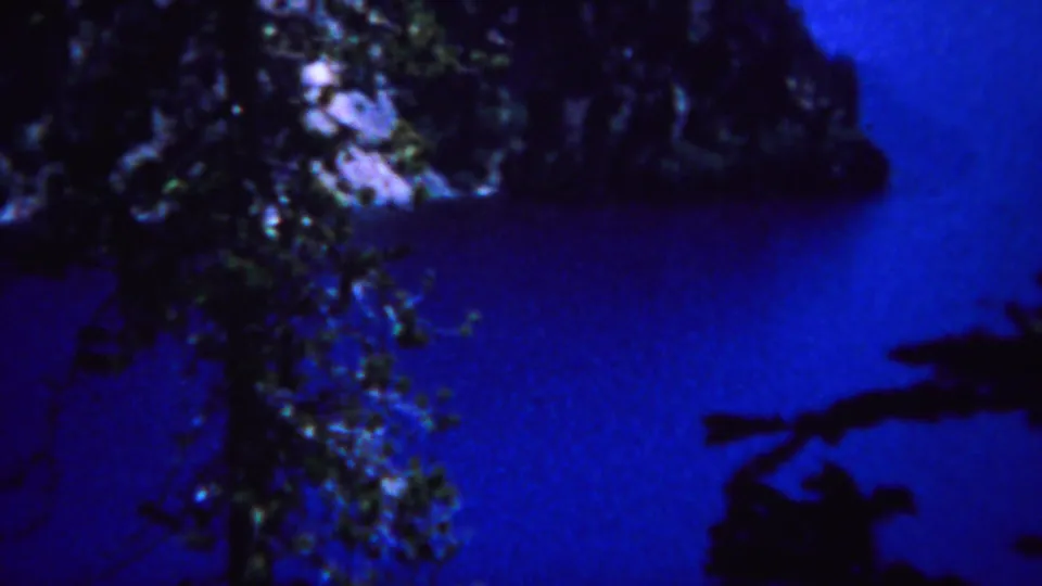 1967 Yellowstone home-movie frame with dark evergreens and shoreline against a vivid blue lake surface.