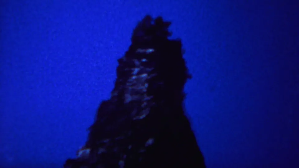 A narrow Yellowstone rock spire stands in dark silhouette against vivid blue 8mm color.
