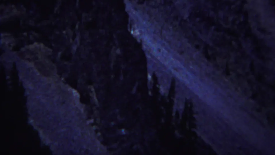 1967 Yellowstone home-movie frame showing shadowed rock faces and dark evergreen shapes under a blue low-light cast.