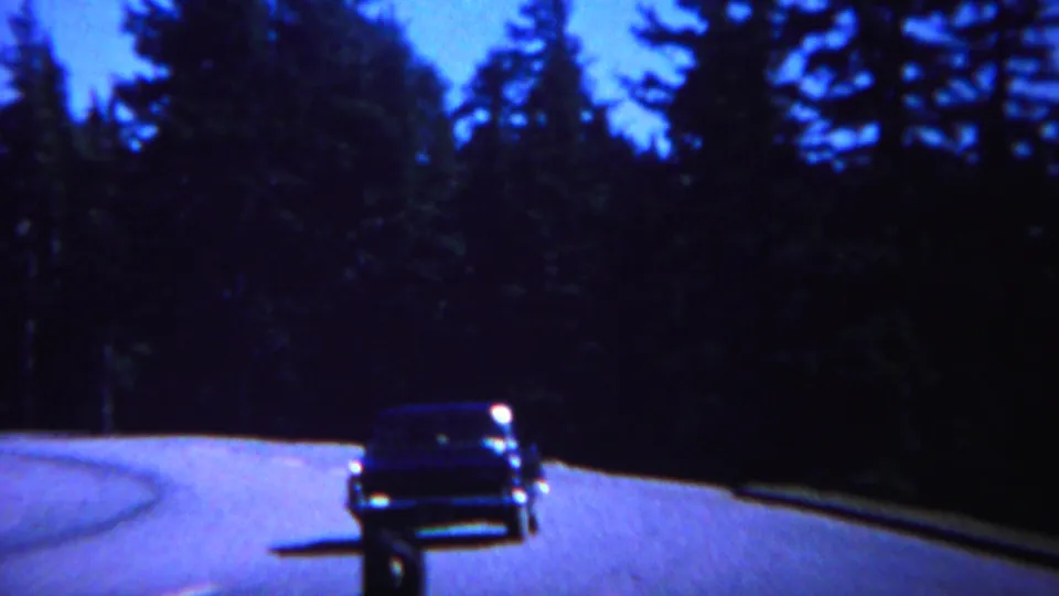 1967 Yellowstone 8mm frame showing a dark classic car on a winding forest road as a black bear moves near its path.
