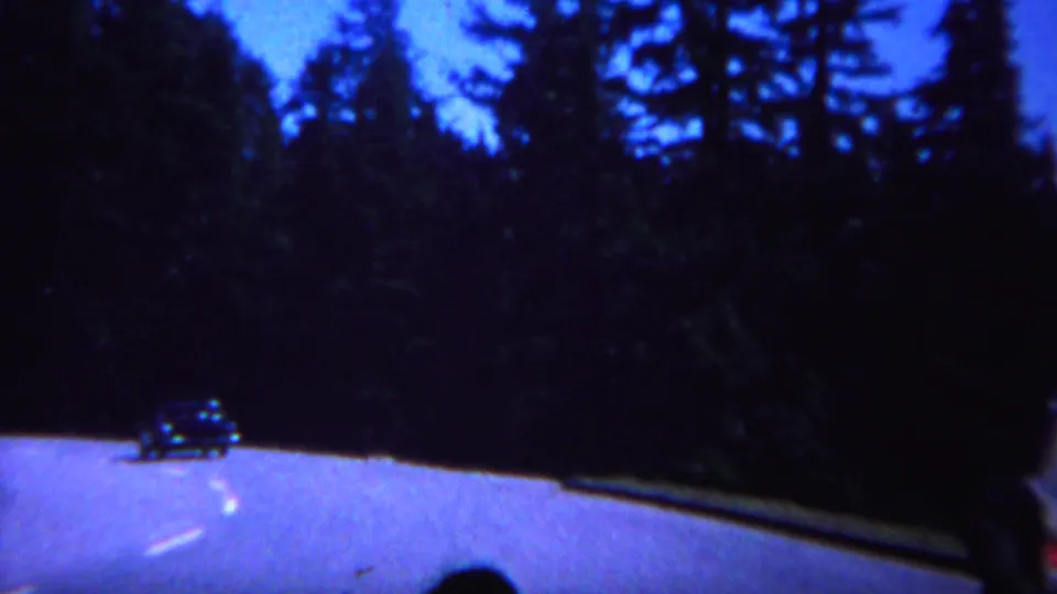 1967 Yellowstone road footage with a distant classic car, dark conifers, and a partial in-car figure at the bottom of frame.