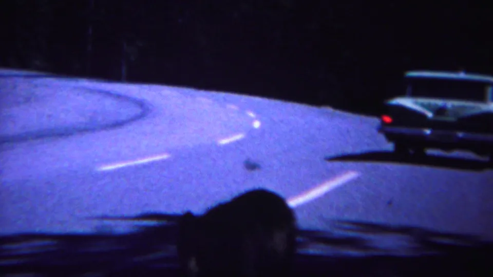 A classic car passes close to the camera vehicle on a winding Yellowstone forest road, with a dark passenger silhouette in front.