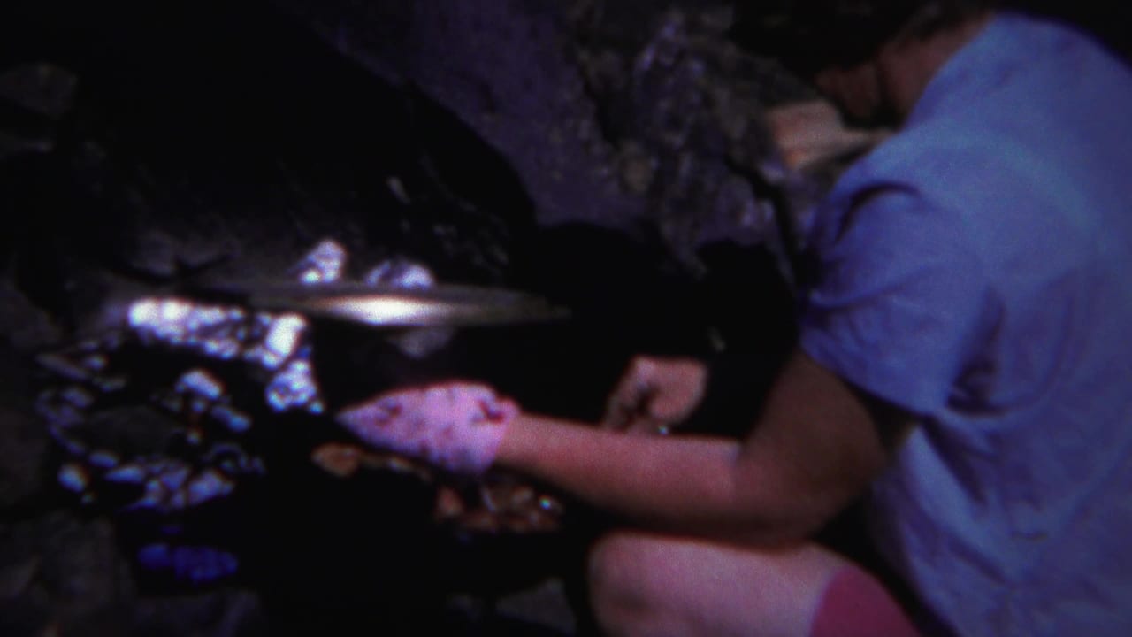 Woman crouching beside a rocky campfire while tending food near firewood.