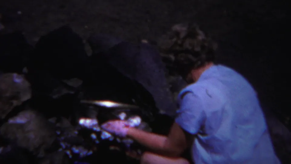 Seen from behind in 1967 Yellowstone footage, the woman crouches close to the fire while tending campsite food.