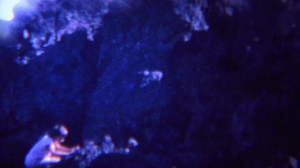 A child crouches at the base of a dark rock wall in faded blue-purple 8mm footage from the Yellowstone-area travel scene.