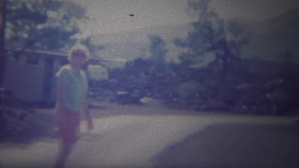 Adult woman near a paved Yellowstone walkway, with a tree-lined background and low building visible through faded 8mm color.