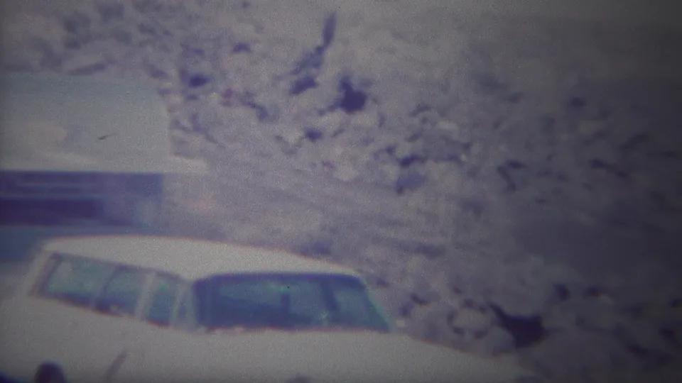 A parked vintage car appears near rocky Yellowstone terrain and a low structure at the end of the home-movie shot.