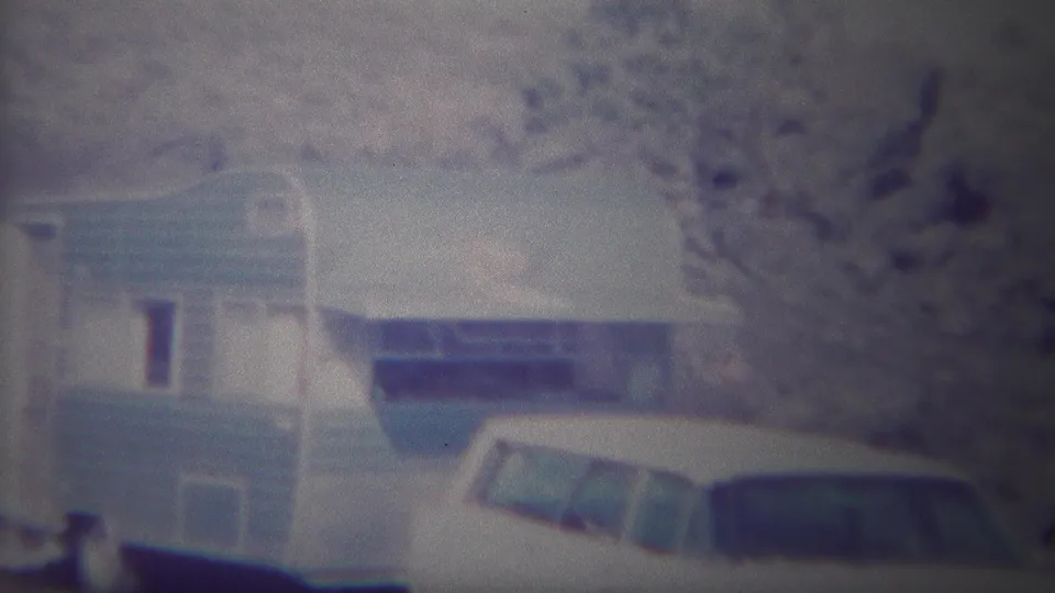 Grainy 1967 Yellowstone frame with a light blue camper trailer and white station wagon parked below trees and hillside.