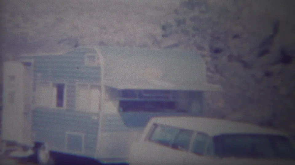 Archival 8mm frame of a camper trailer and station wagon at a Yellowstone campsite, with trees and dry hillside in the background.