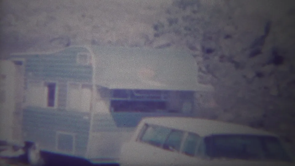 1967 Yellowstone home-movie frame showing a blue-and-white camping trailer with striped siding, sparse trees, and a white vehicle partly visible in the foreground.