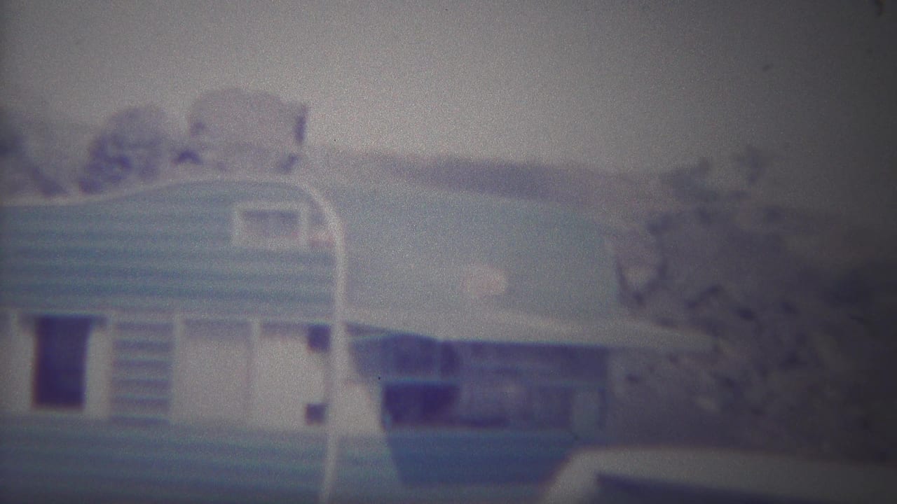 1967 Yellowstone frame with the camper trailer centered, showing its rounded roof, blue stripe, side door area, and trees on a rocky hillside.