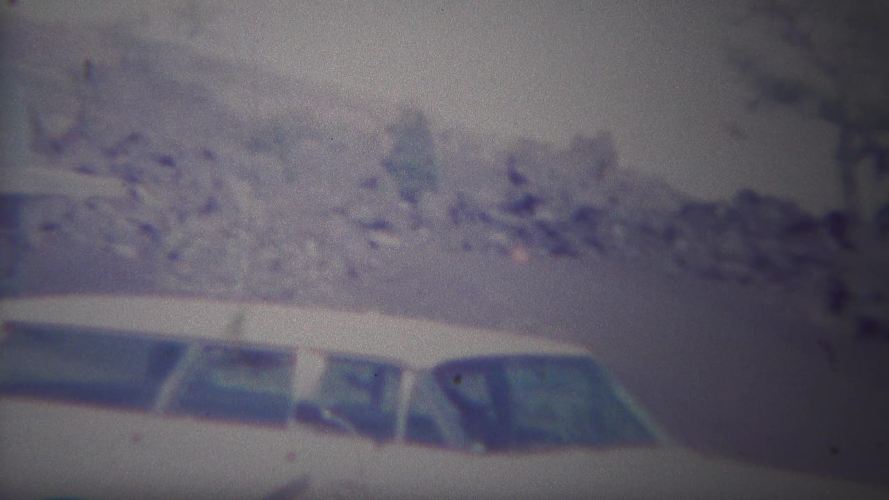 1967 Yellowstone-area home-movie frame with white cars in front of scrubby brush, pale rock, and sparse trees.
