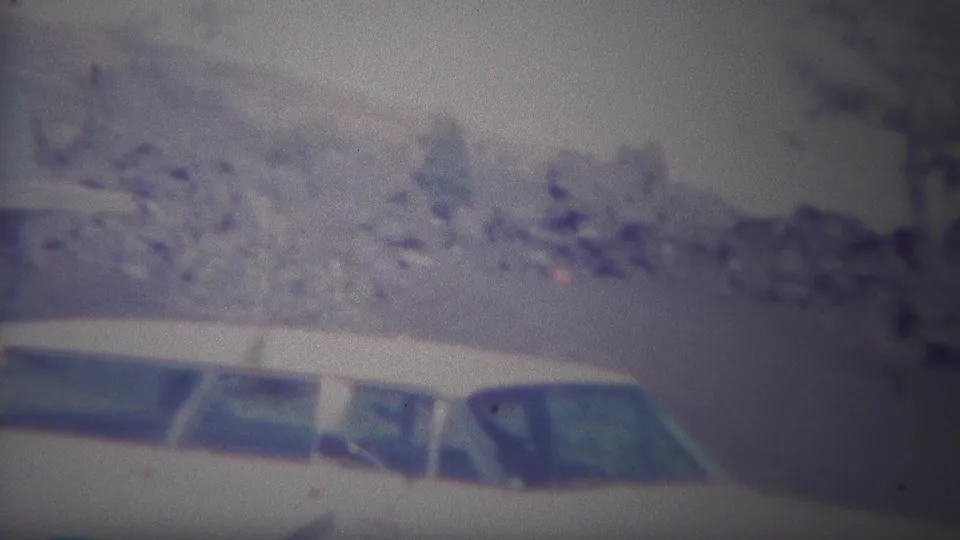 1967 Yellowstone-area home-movie frame with white cars in front of scrubby brush, pale rock, and sparse trees.