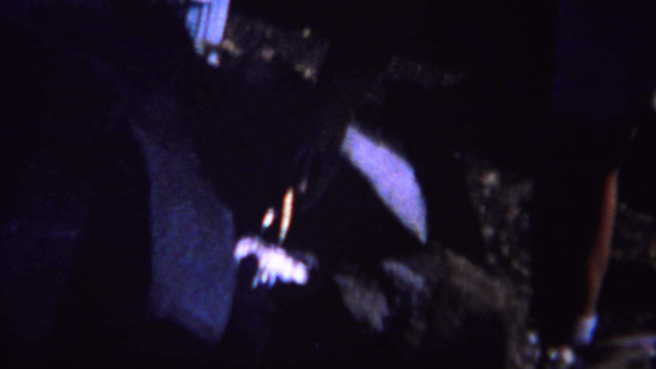 Dark, soft-focused opening frame with partial foreground shapes and outdoor shadows.