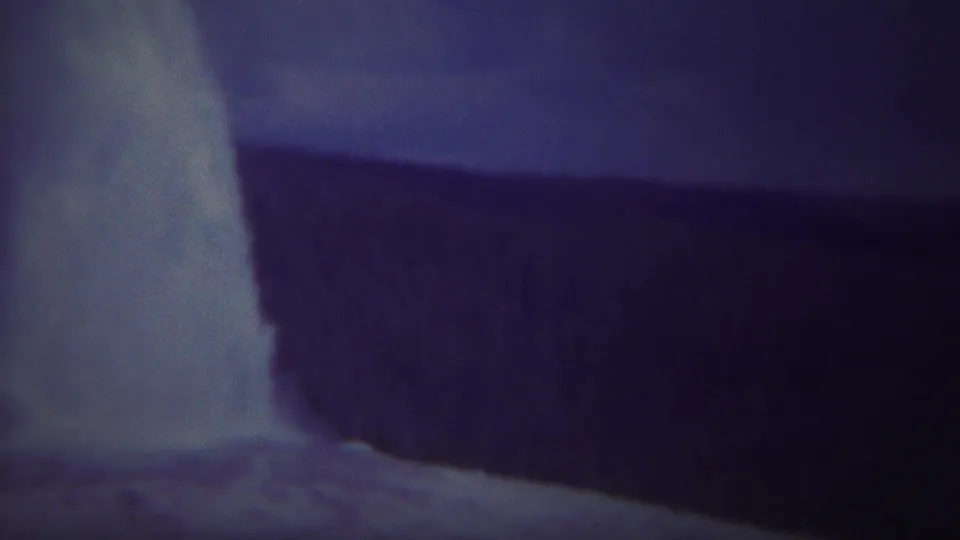 Old Faithful erupts in a tall pale plume beside the dark Yellowstone tree line in aged 1967 8mm footage.