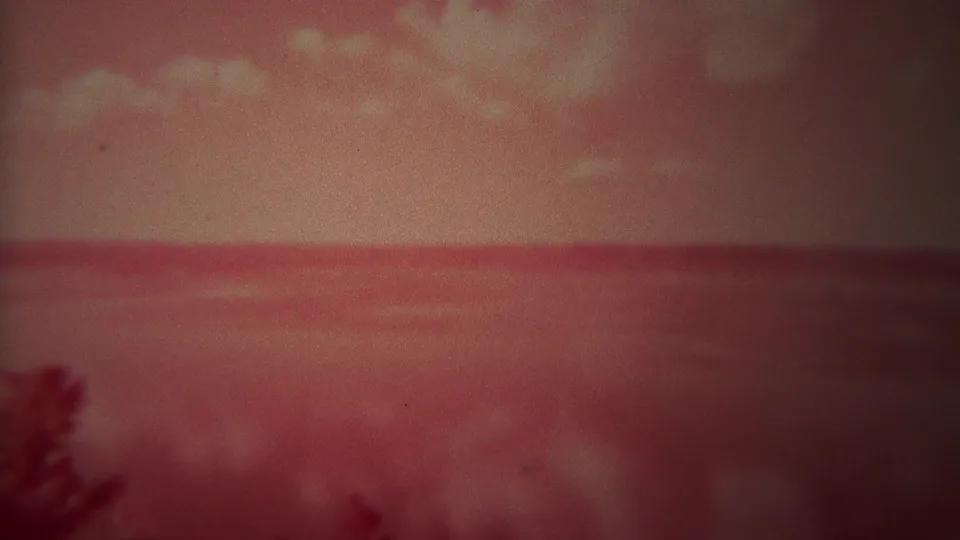 1967 Yellowstone 8mm frame of a calm red-tinted water surface, clouded sky, and a soft dark tree silhouette in the foreground.