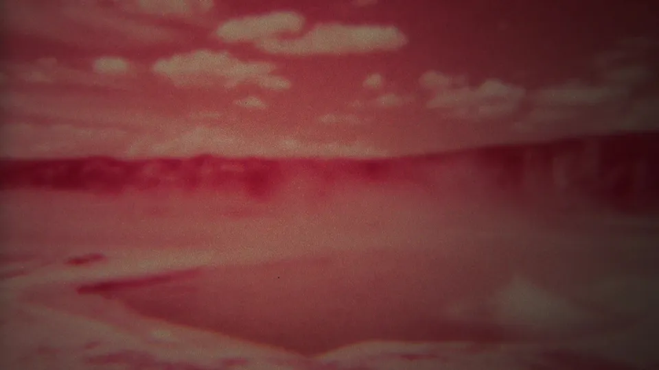1967 8mm Yellowstone frame showing cloudy sky above a red geothermal basin with steam and soft foreground water.