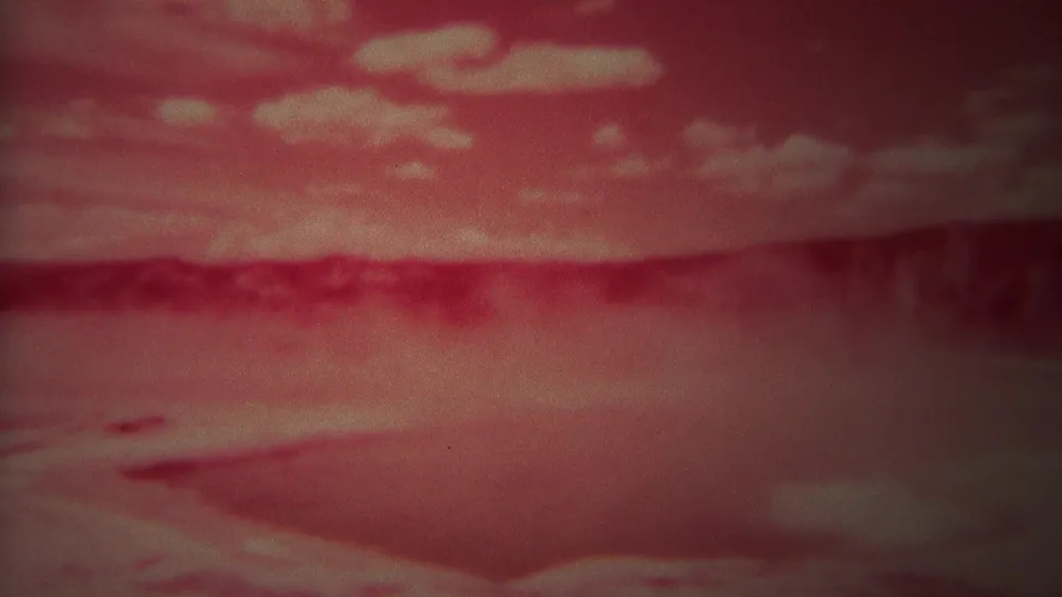 1967 8mm Yellowstone view of a red geothermal basin with pale clouds, steam haze, and a dark ridge line.