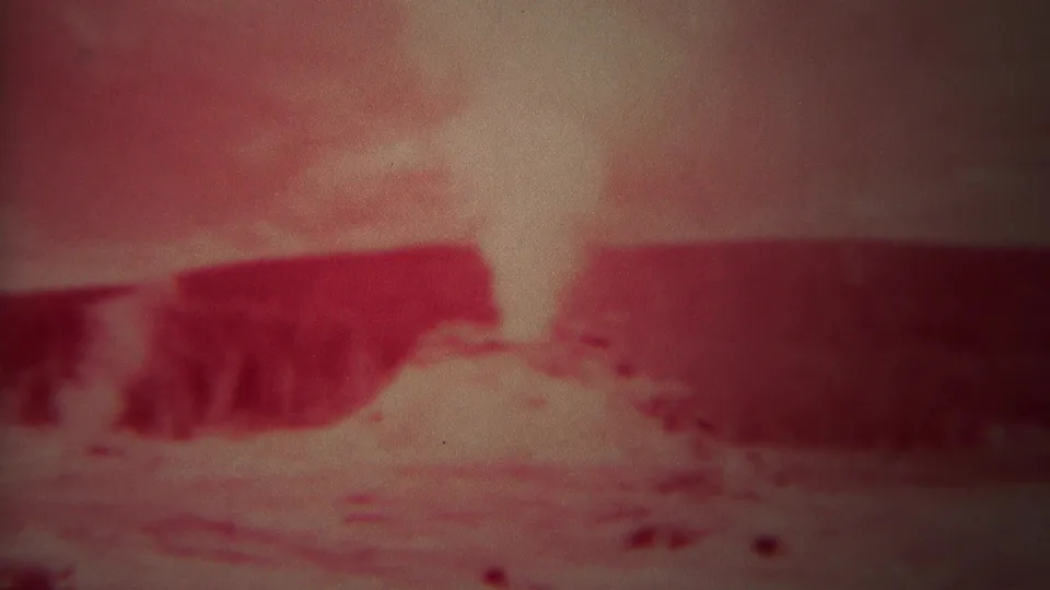 1967 Yellowstone 8mm frame showing a bright pale steam or spray plume above reddish terrain and a dark ridge, with grain and color fading.