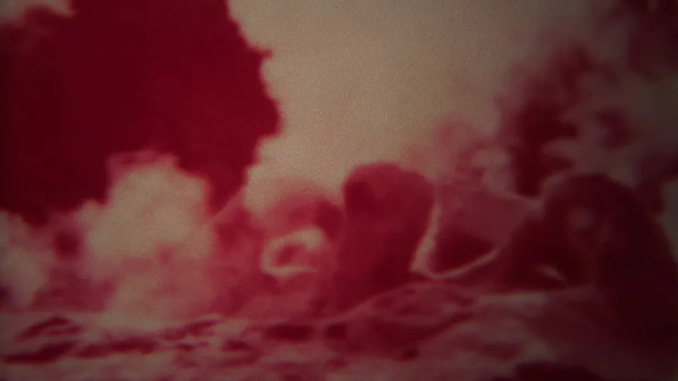 1967 Yellowstone home-movie frame with steam drifting across dark rocks and a rounded reddish geyser formation.