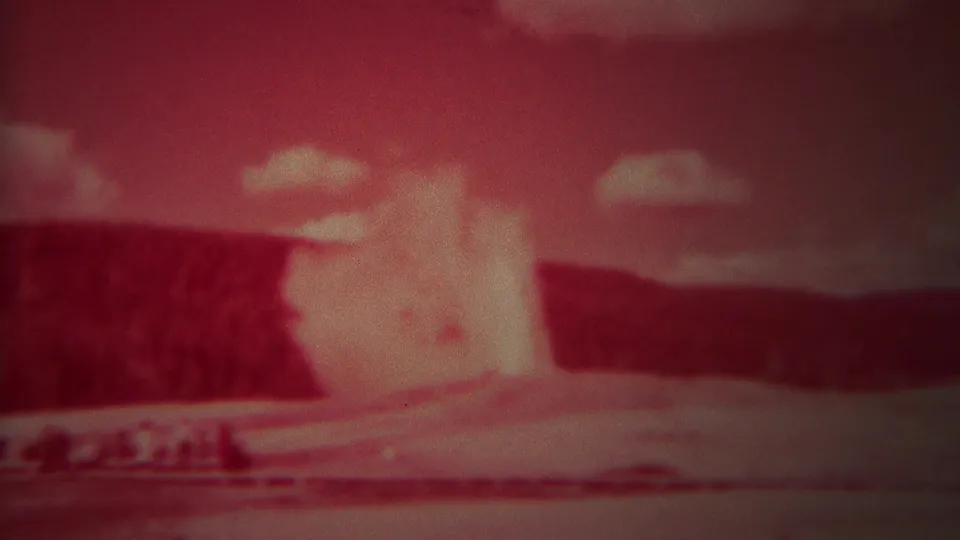 Red-tinted 1967 Yellowstone 8mm frame with Old Faithful erupting in the distance beyond a low viewing area.
