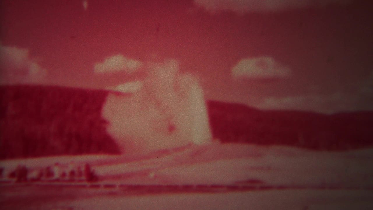 Red-tinted 8mm frame of a geyser plume rising behind a distant viewing area.