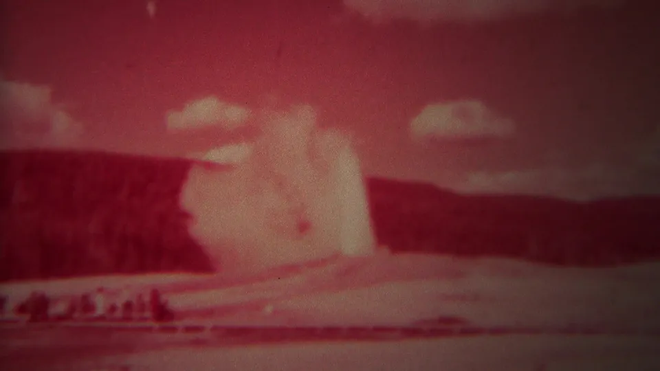 1967 Yellowstone home-movie frame showing Old Faithful erupting with visitors faintly visible near the viewing area.
