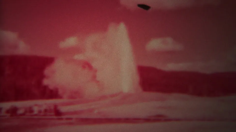 1967 Yellowstone frame of Old Faithful erupting, with distant visitors below and a dark mark in the film near the top edge.