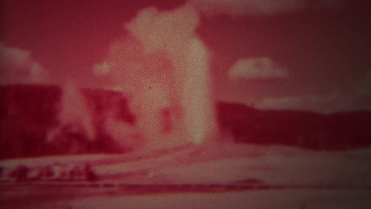 1967 Yellowstone home-movie frame showing Old Faithful erupting in a strong red tint with distant hills and clouds.