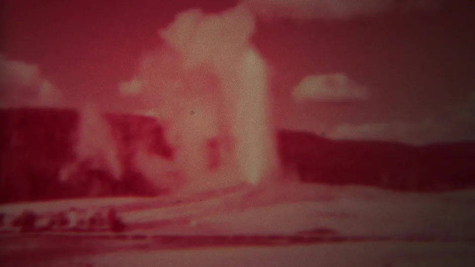 1967 Yellowstone home-movie frame showing Old Faithful erupting in a strong red tint with distant hills and clouds.