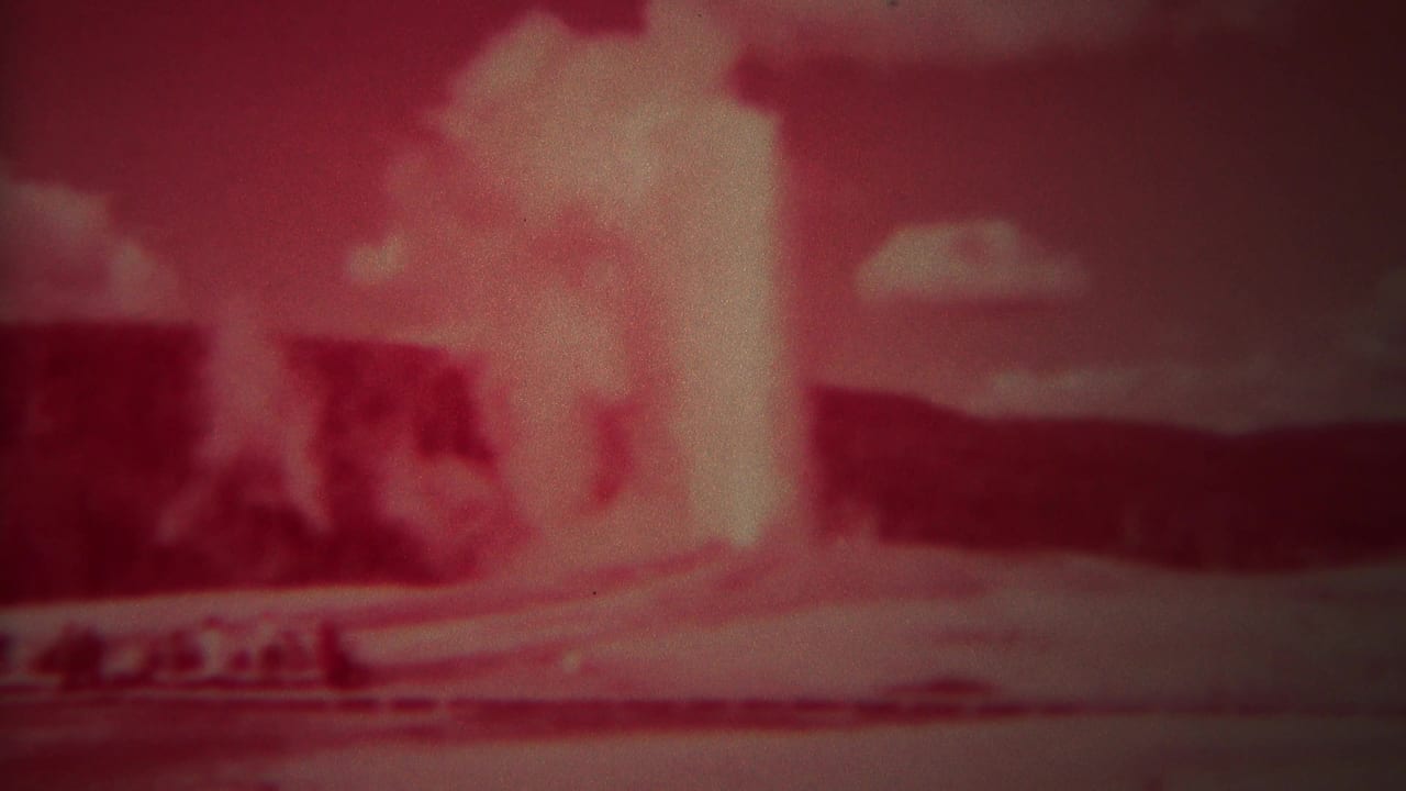 Old Faithful eruption fills the center of a red-tinted frame with dark hills behind.