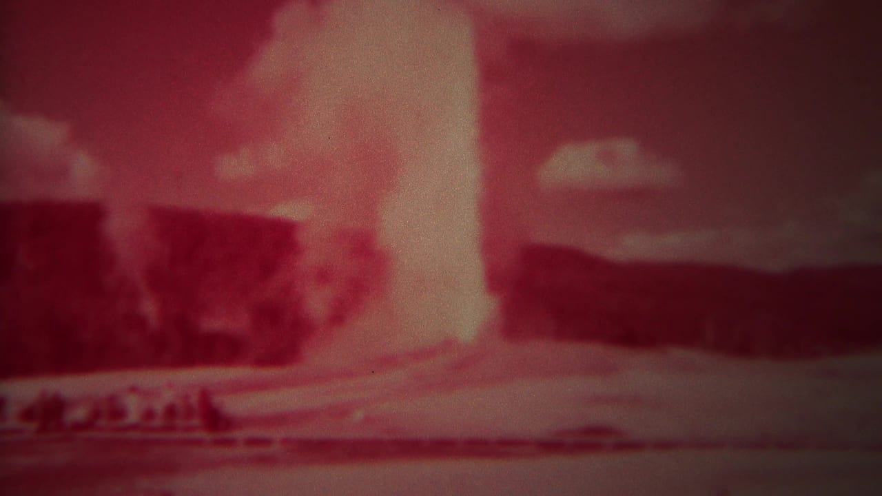 Red-tinted geyser plume rising behind a low foreground ridge and distant hills.