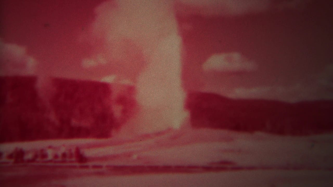 Red-tinted geyser eruption continuing beside dark hills and a bright foreground area.