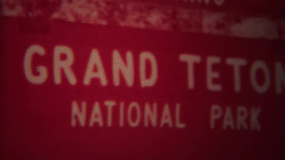 1967 8mm view of the Grand Teton National Park sign with cropped white lettering, reddish color shift, and soft focus.