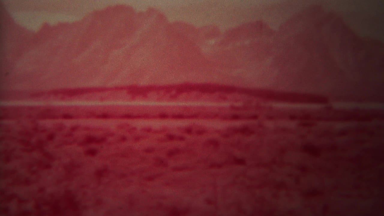 Red landscape with scrubland foreground, dark ridge line, and hazy mountains.