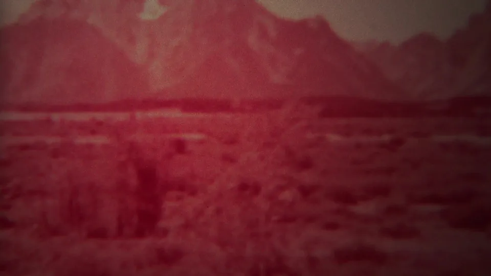 Vintage 8mm Yellowstone, Wyoming landscape with a red color cast, grainy brush foreground, pale midground, and distant mountains.